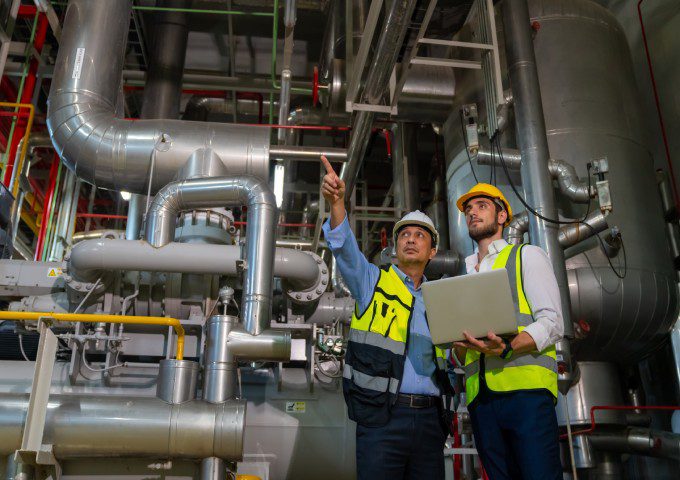 Two men in hard hats and vests are standing next to pipes.