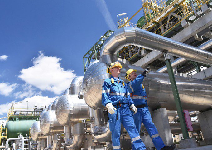 Two men in blue suits and hard hats standing next to a pipeline.