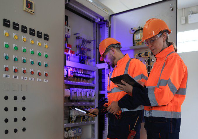 Two men in orange jackets and hard hats are looking at a control panel.